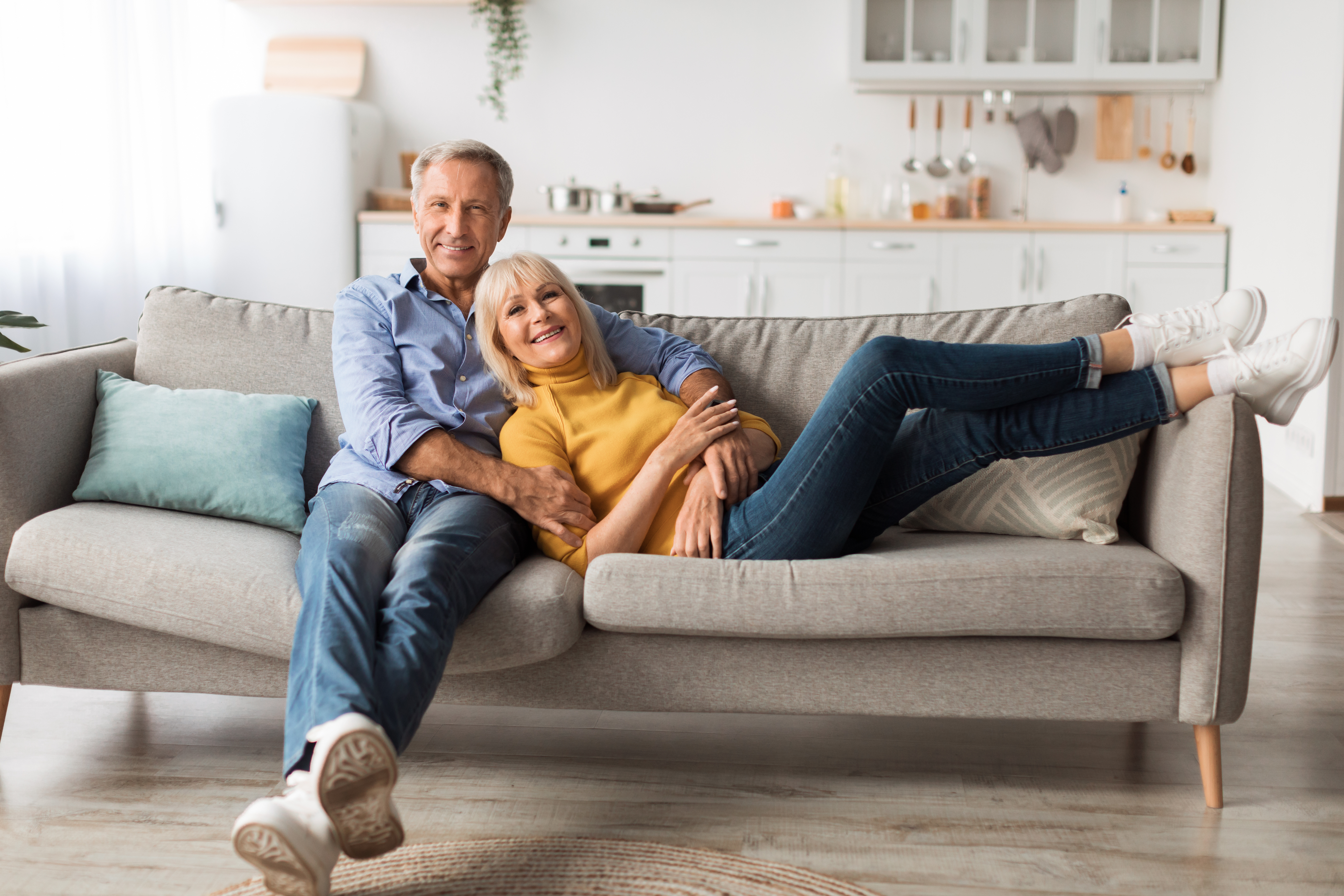 Mature Spouses Hugging Sitting On Sofa Together At Home, Smiling To Camera. Happy Senior Husband And Wife Embracing Relaxing On Couch On Weekend. Retirement Lifestyle And Happiness In Marriage